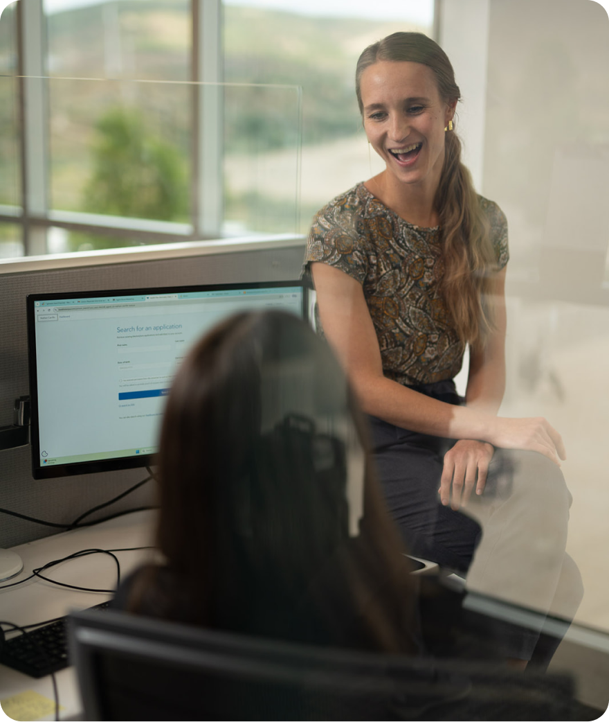 Two women talking in an office, one sitting on other's desk